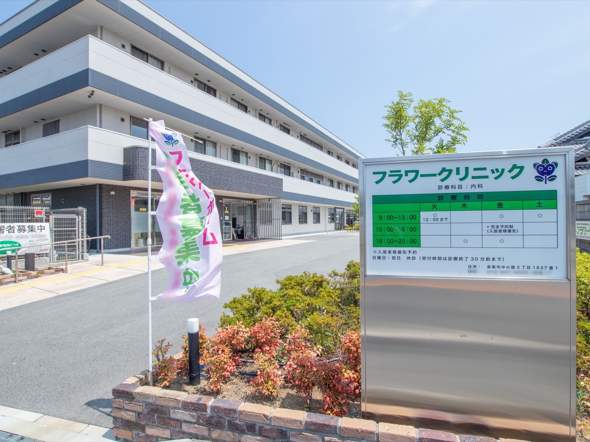 施設建物の手前にレンガで囲った植え込みがあり、植物が植えられている。その横にはクリニックの看板が掲げられている。