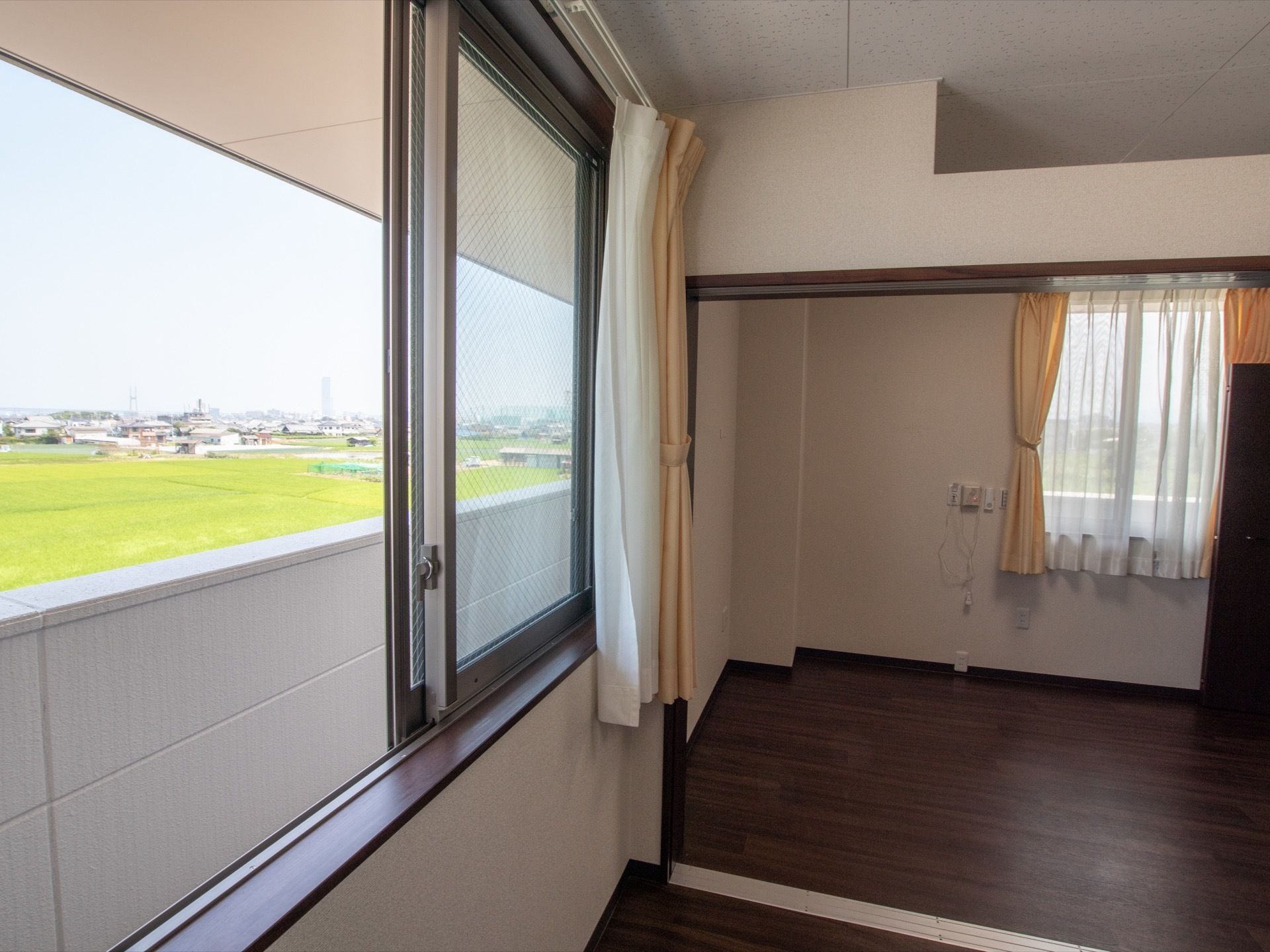 開け放された居室の窓からは田んぼや近隣の住宅が見える。部屋は二間続きとなっていて、奥の部屋にも窓がある。