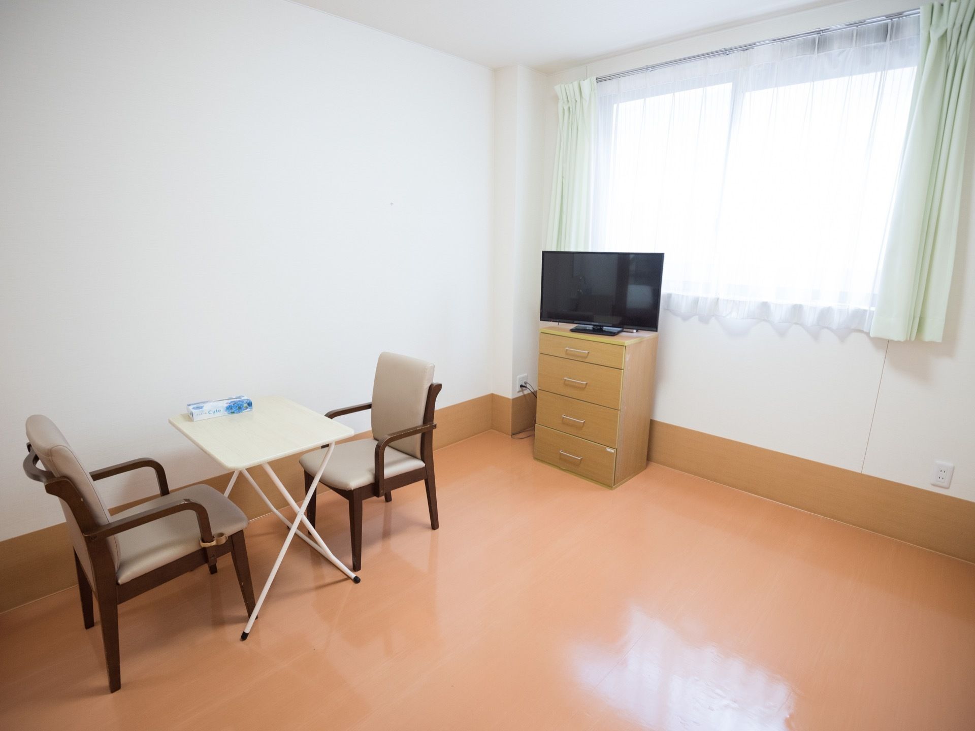 薄いオレンジ色の床が敷かれた居室があり、テーブルセットがある。窓際にはチェストがあり、その上にテレビがのっている。