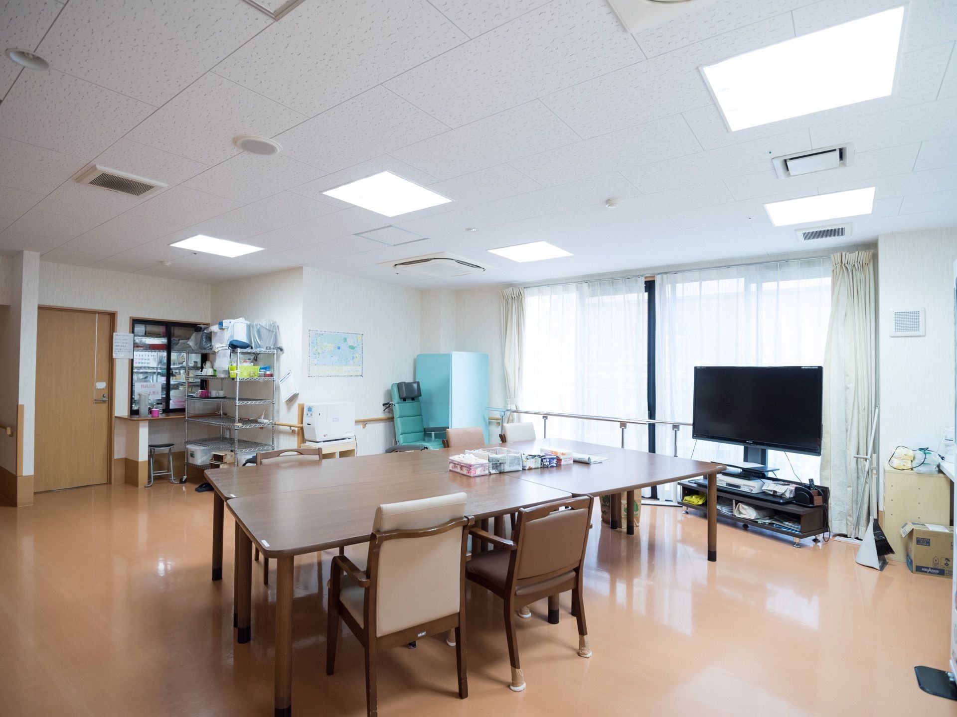 掃き出し窓のある明るい食堂がある。中央にはテーブルや椅子があり、窓際には大型のテレビがある。