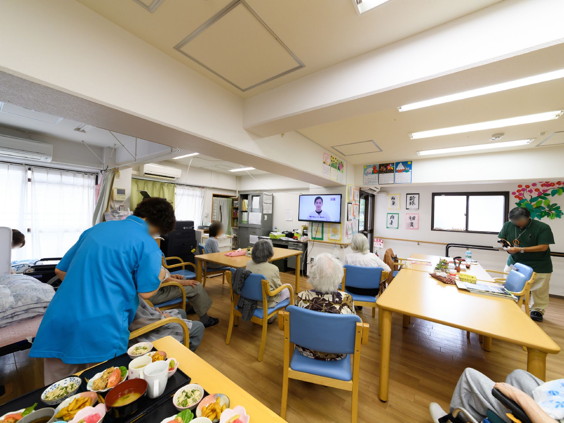 食堂ではたくさんの入居者様が椅子に座り、テレビをみている。テレビでは女性が一人うつっている。女性スタッフがお世話をしている。