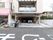 施設は道路に面してたてられており、左には司法書士事務所が入ったビル、右には一般の民家がたっている。道路には、とび出し注意と表記されている。
