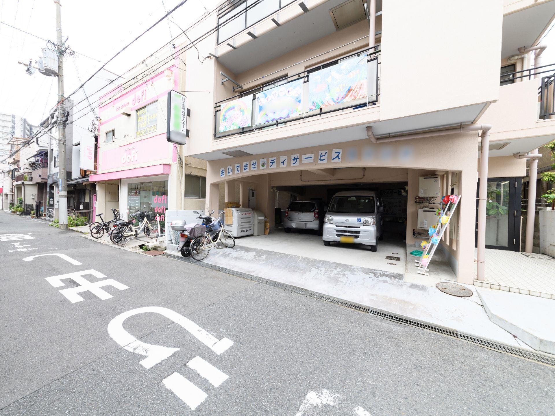 施設の1階には駐車スペースがあり、自動車が2台とまっている。自転車とバイクも1台ずつとまっている。施設名の看板が設置されている。