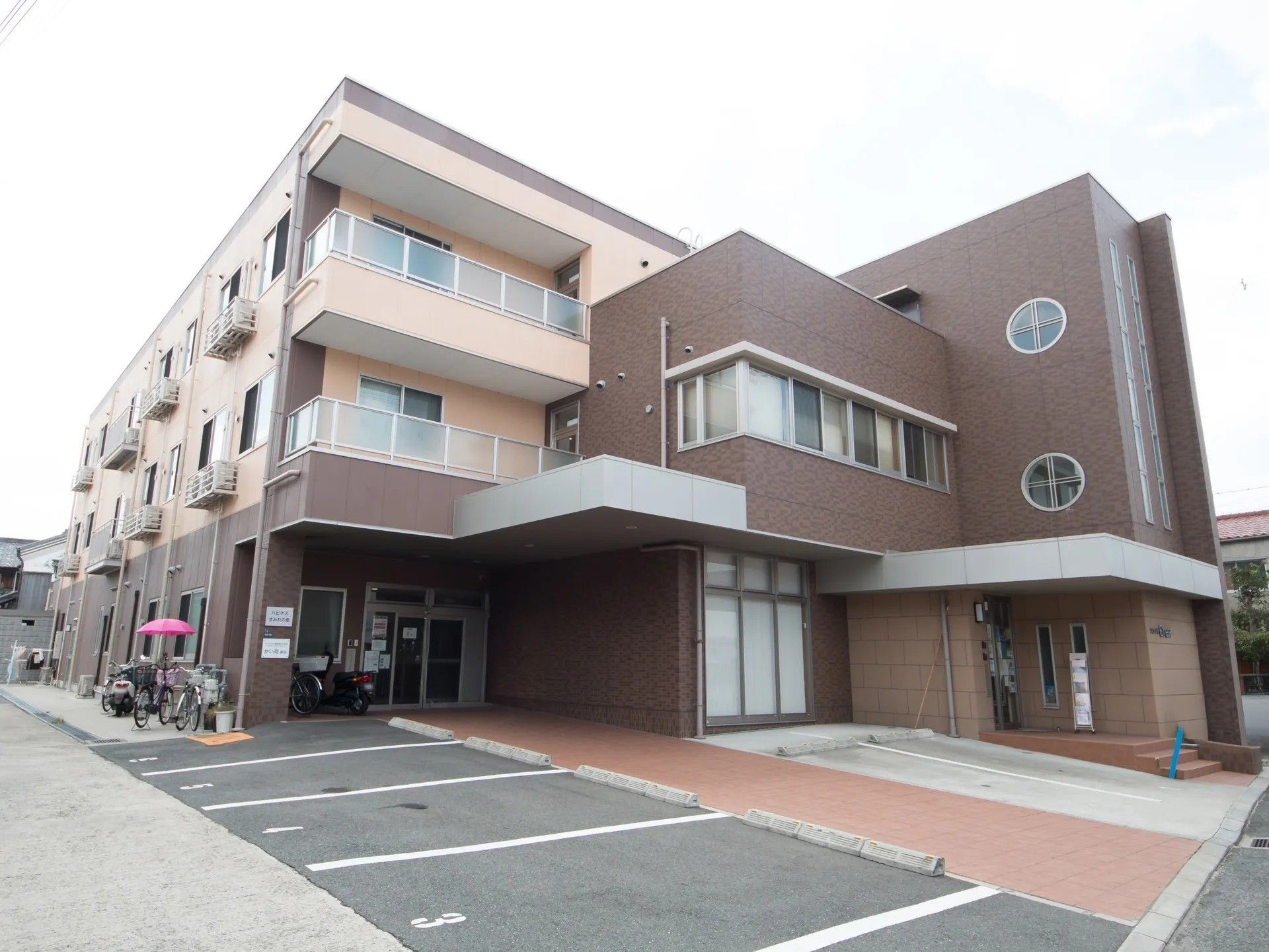 やや斜めの角度から施設建物を見上げたところ。駐車用のスペースが設けられており、その間にブロック舗装のされた歩行者用通路がある。