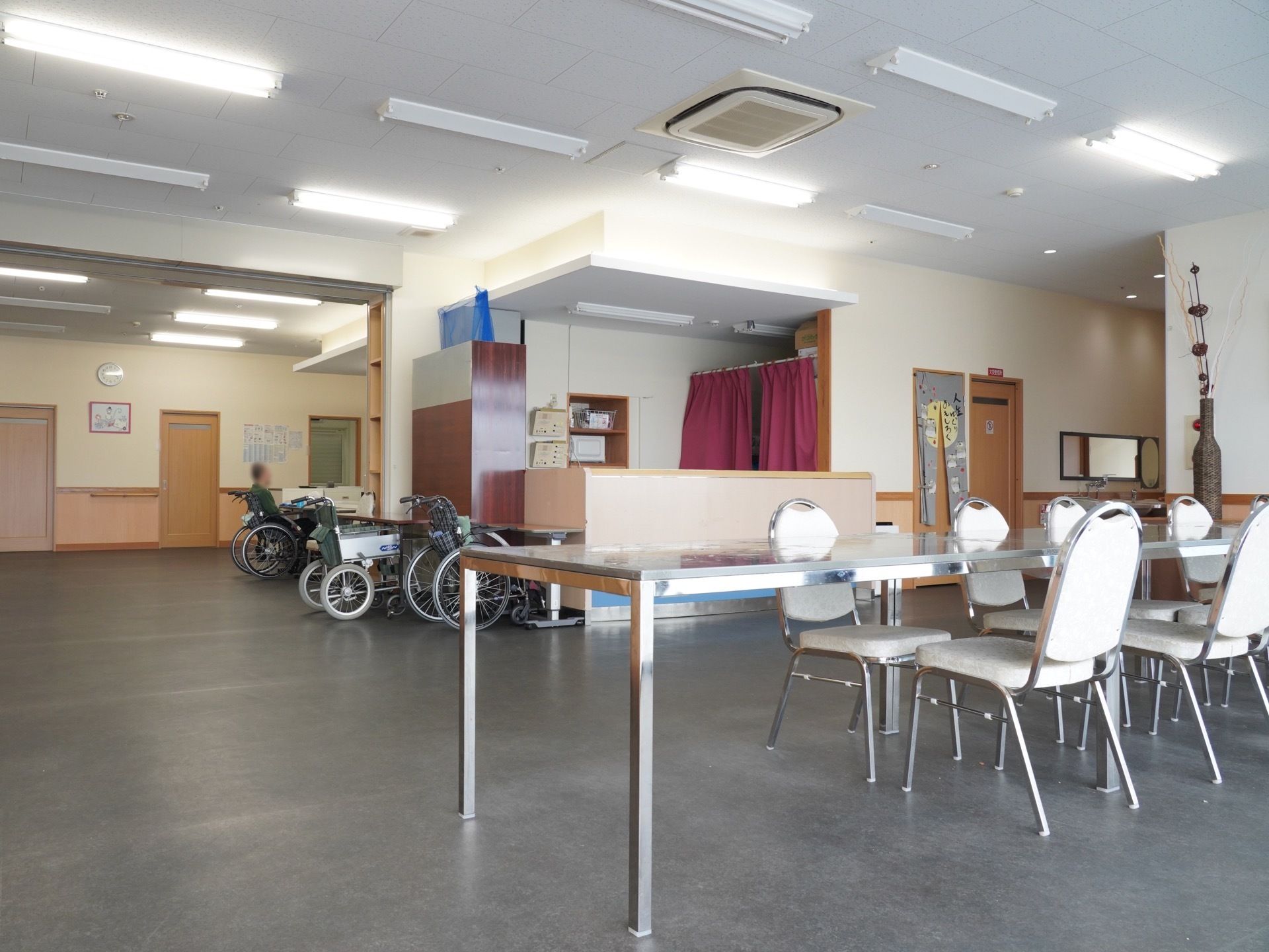 広い部屋に長机と、背もたれ付きの椅子が並んでいる。奥には車椅子が並べられており、入居者様が一人座っている。
