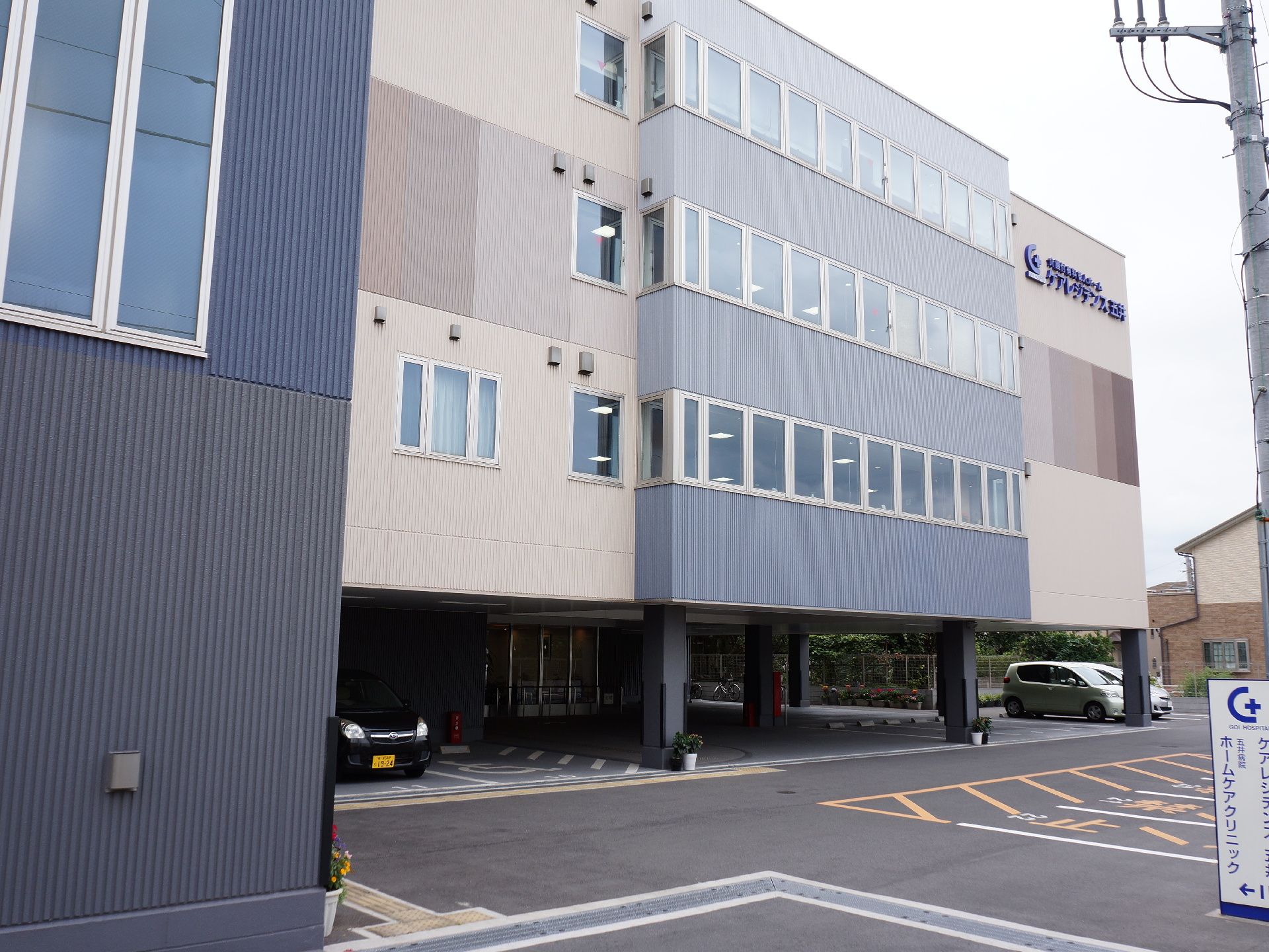 外観のデザインは、シックで落ち着いた雰囲気となっている。電柱の前には白い看板があり、施設名とクリニック名が2つ書かれている。