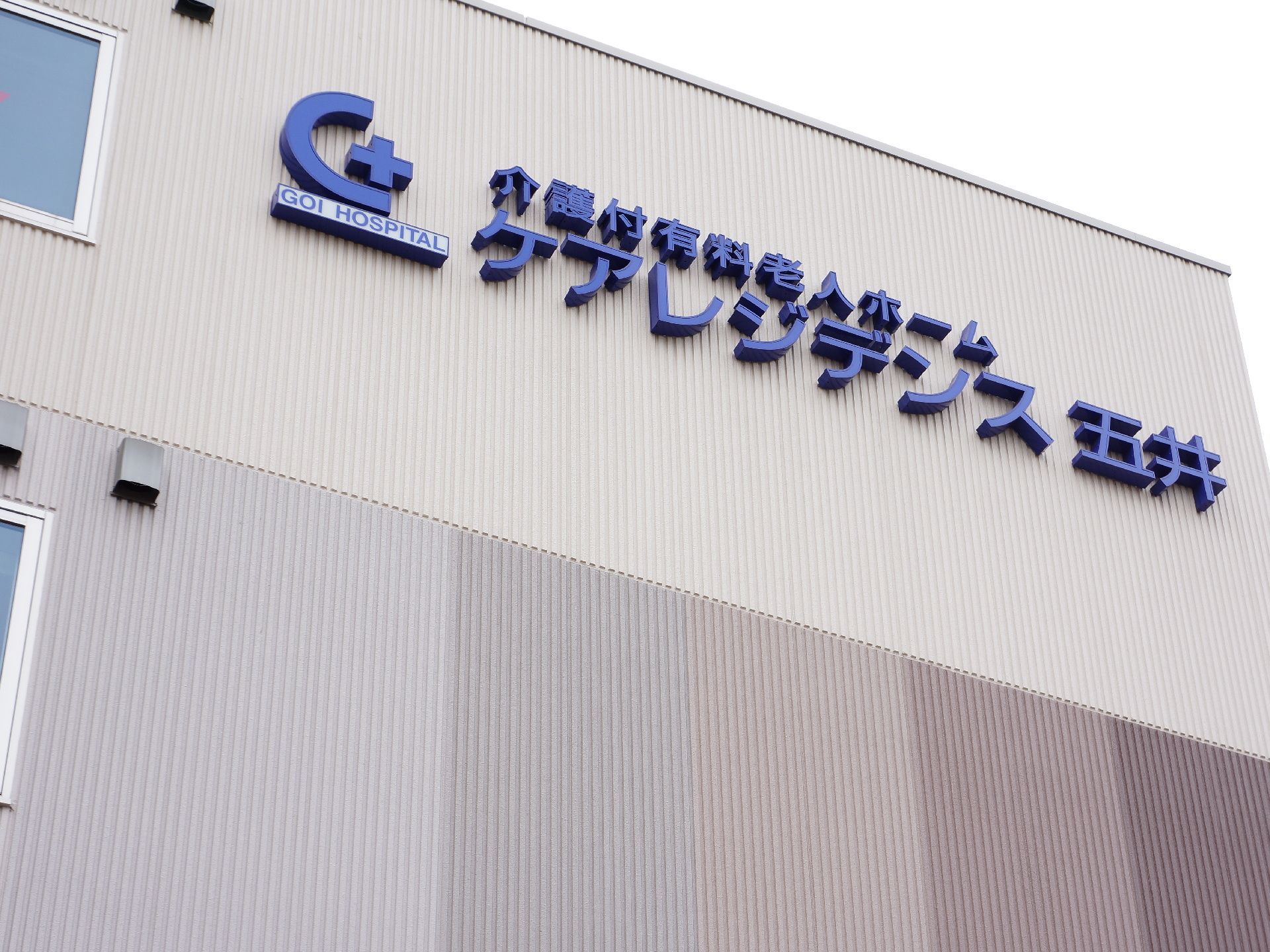 ベージュ色の壁に、施設名が大きな紫色の文字で書いてある。その横には、アルファベットのGの形をモチーフにしたマークがある。