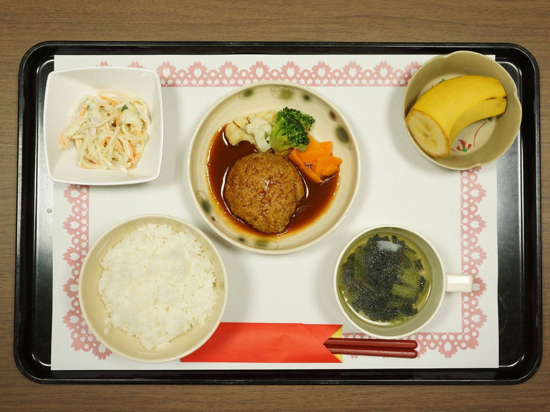黒いお盆の上にクロスを敷いて食事を置いている。白米、スープに、ハンバーグやサラダ、バナナといったものが乗っている。