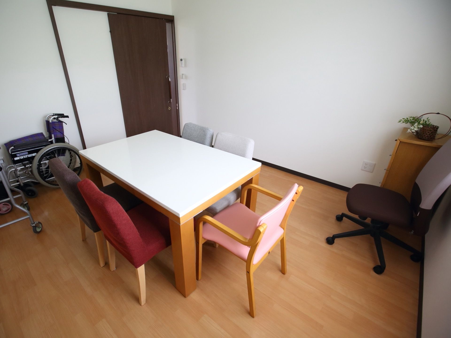 フローリング床の部屋である。木製のテーブルに椅子が４脚セットされている。隣に回転椅子が１脚置かれている。壁際に車椅子がある。