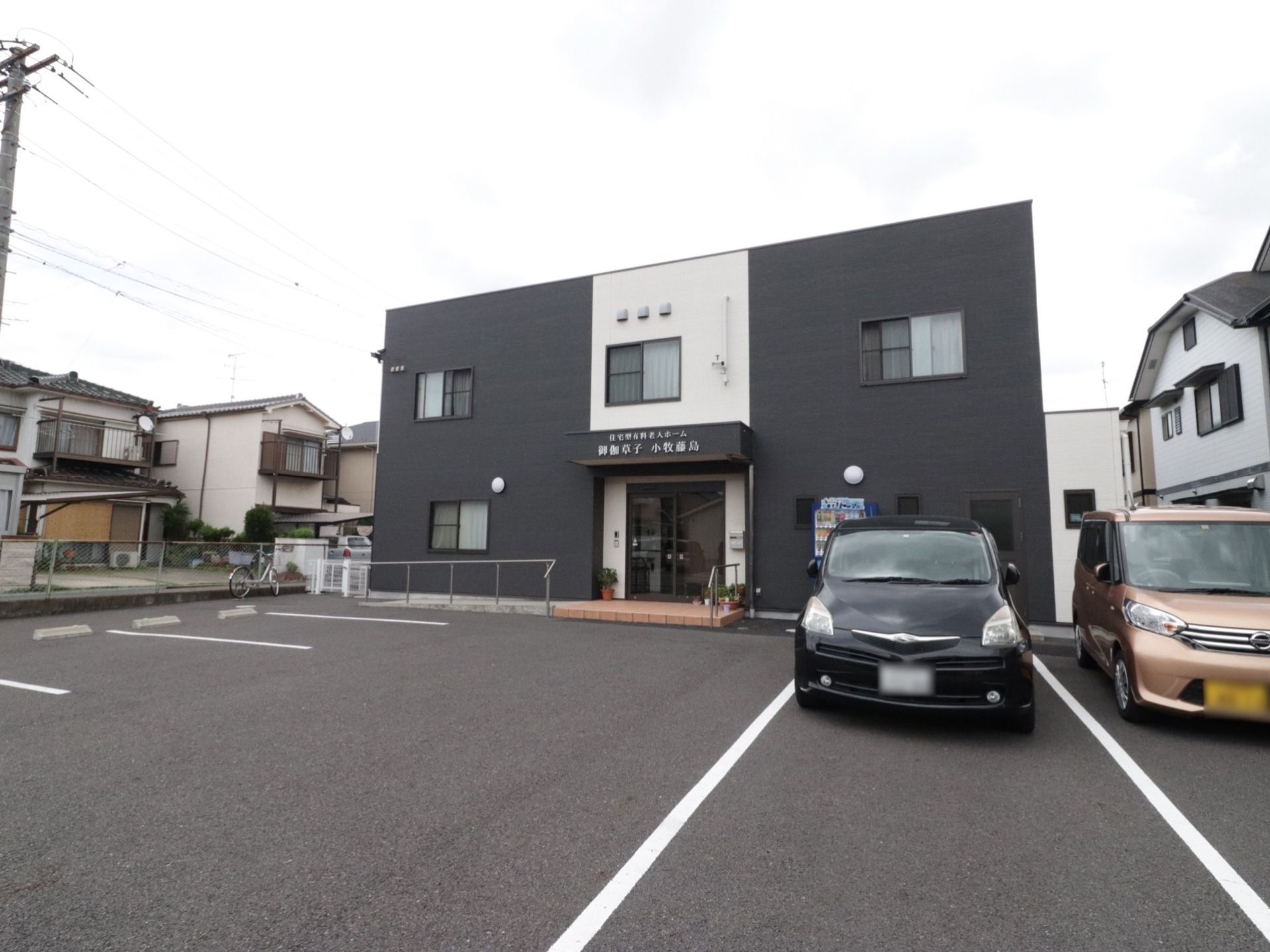 モノトーンで統一された建物になっている。手間には駐車場があり、車が数台停まっている。周辺は閑静な住宅街となっている。