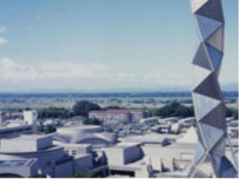 青空の下に町並みが広がっている。手前には巨大なスネークキューブのようなオブジェが、上に向かって伸びるような形で建っている。