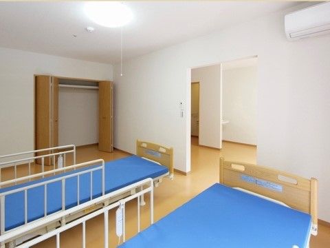 室内には２台の介護用ベッドが置かれ、収納スペースが設けられている。壁は白色基調で、エアコンが設置されている。