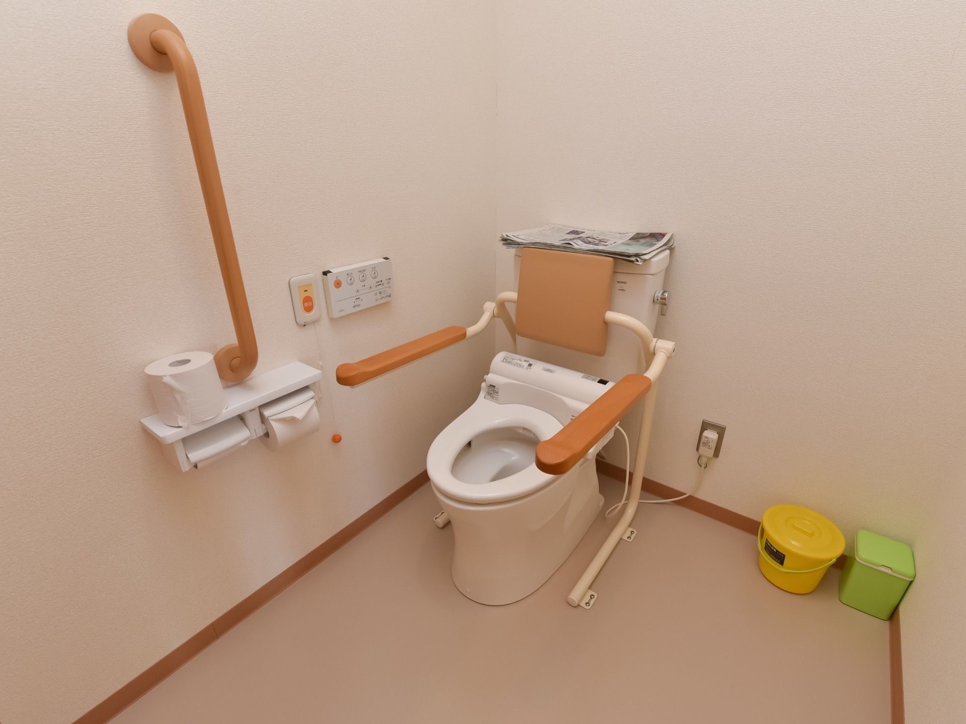 壁には手すりが取り付けられている。便器には背もたれと肘置きがある。小さなゴミ箱が２つある。緊急用ボタンがある。