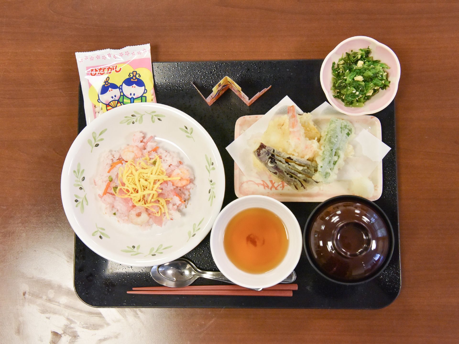 ひな祭りになぞらえた食事が黒いお盆に乗っている。天ぷらに薬味がついており、ちらし寿司に椀物も提供されている。