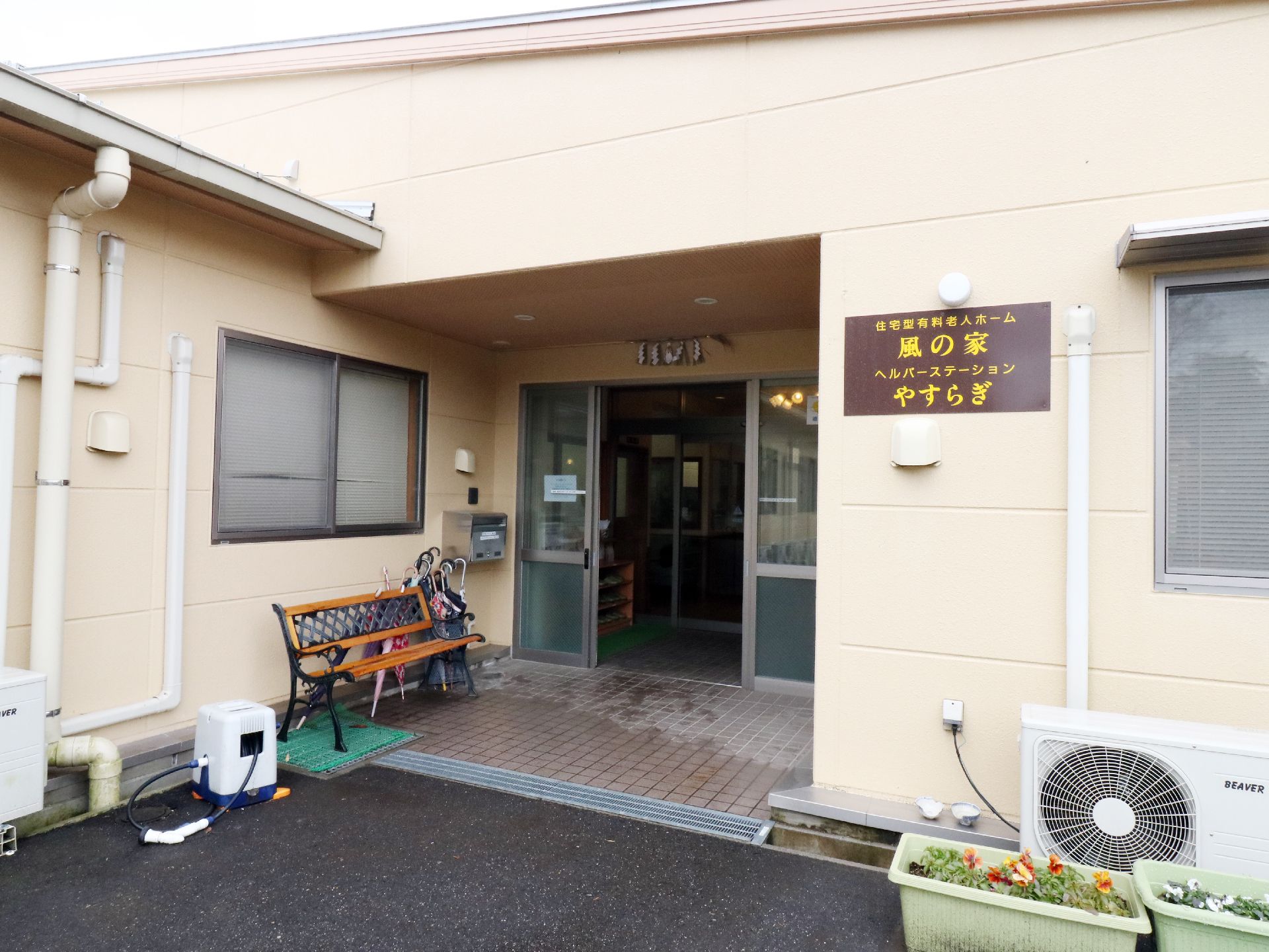 クリーム色の壁に茶色の施設名の入った看板がかけられている。玄関は奥まっており手前にベンチが置かれている。