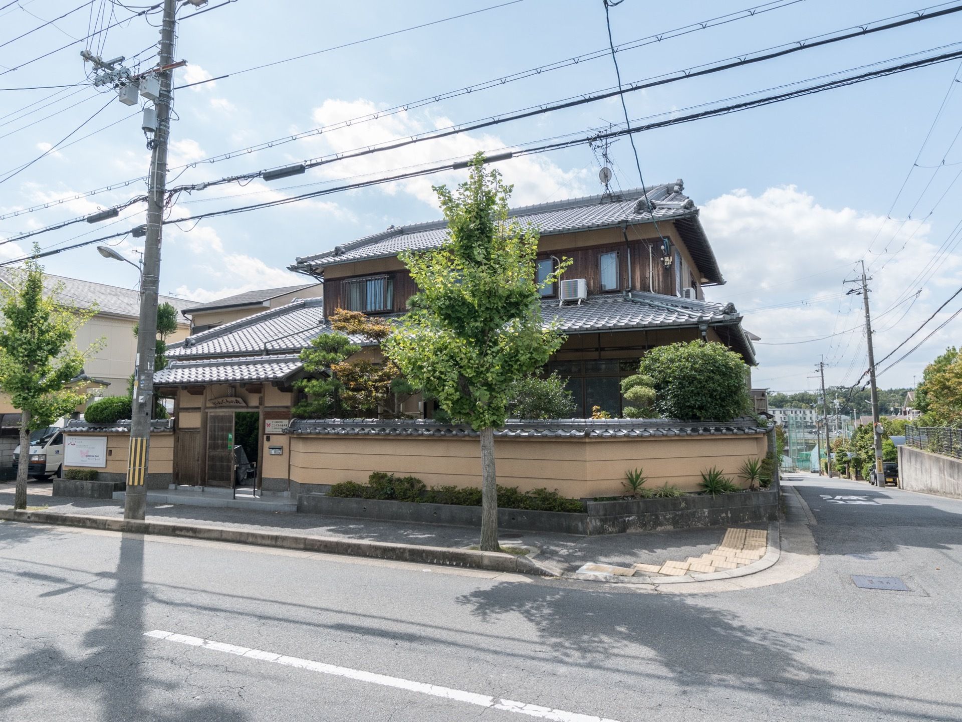 施設は道路に面して建てられている。歩行者通路には大きな木が植えられている。玄関入り口の横には施設の看板が取り付けられている。