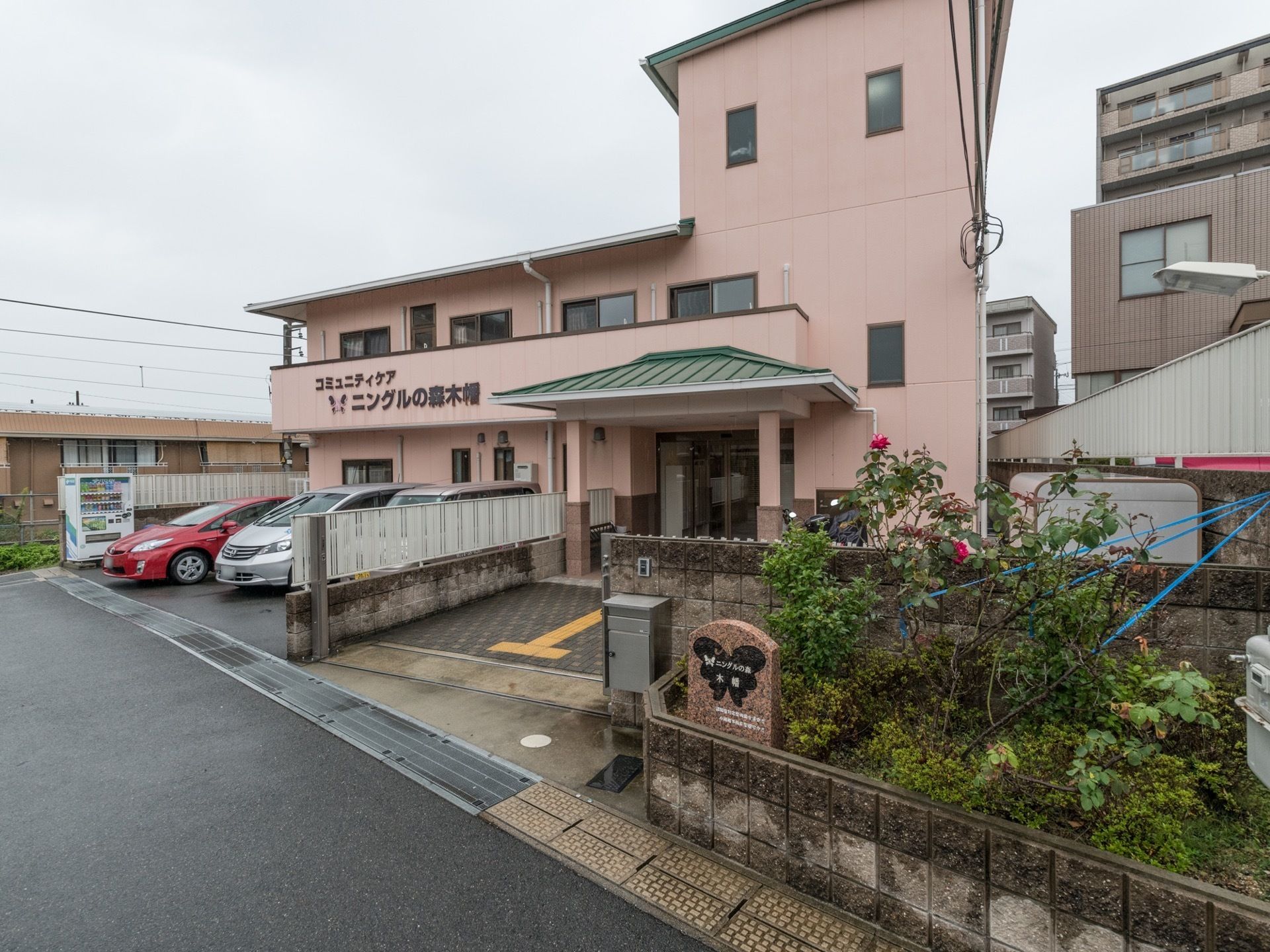 ピンクベージュの外壁に緑色の屋根の建物である。建物前に三台分の駐車場を備えており、自動販売機が設置されている。