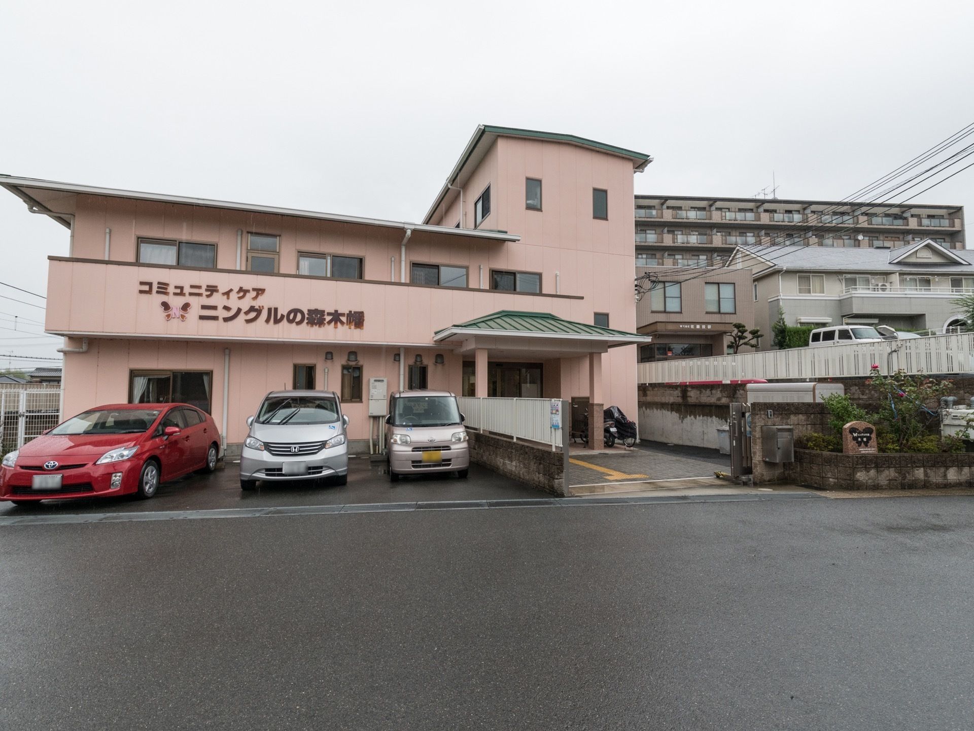 施設は淡いピンク色の建物で、右側は3階、中央から左側は2階建てになっている。前面に駐車スペースを設けている。