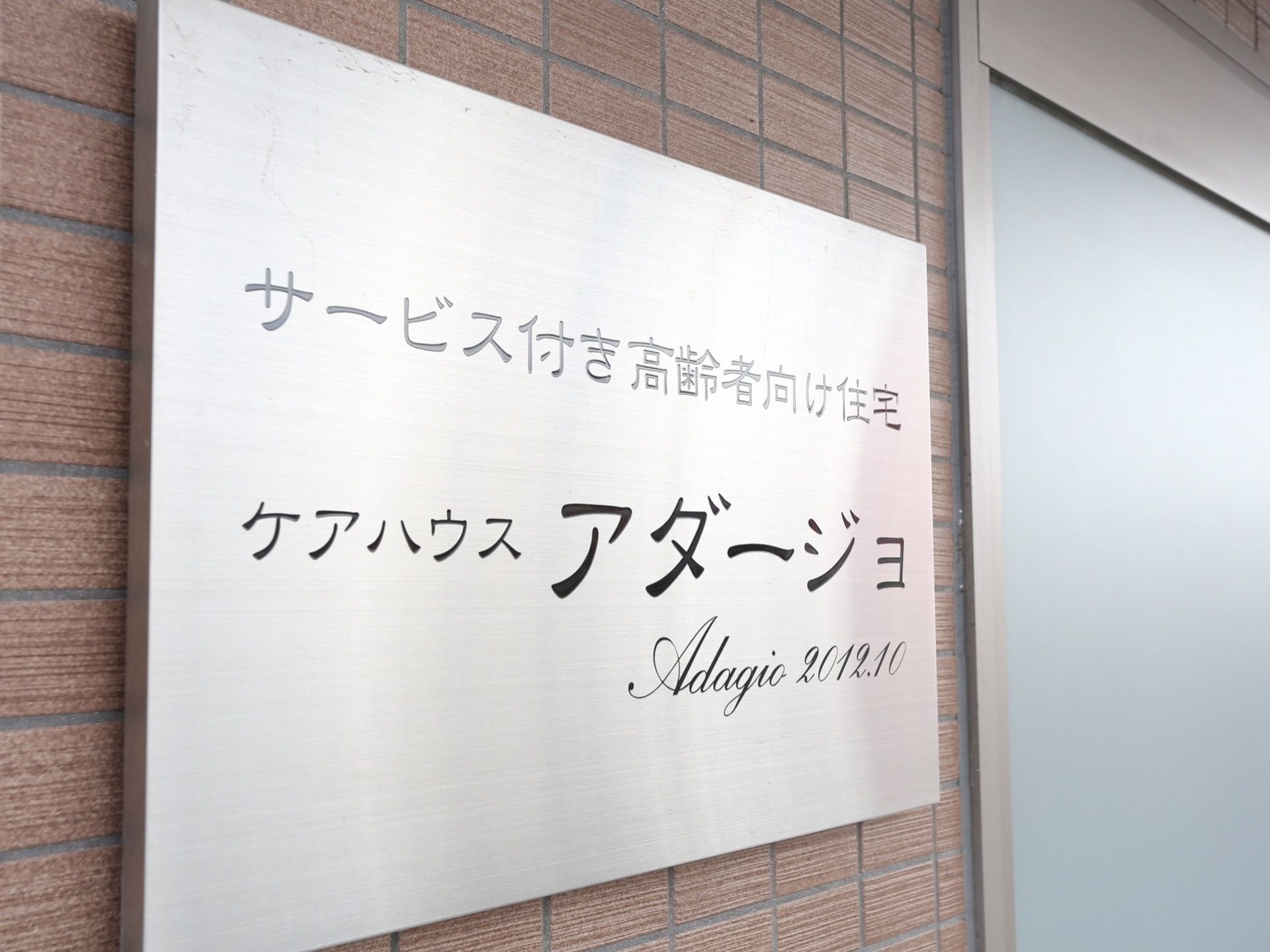 玄関の外は、茶色い外壁となっている。壁には、シルバーのプレートに黒い文字で施設名の書かれた表札が設置されている。