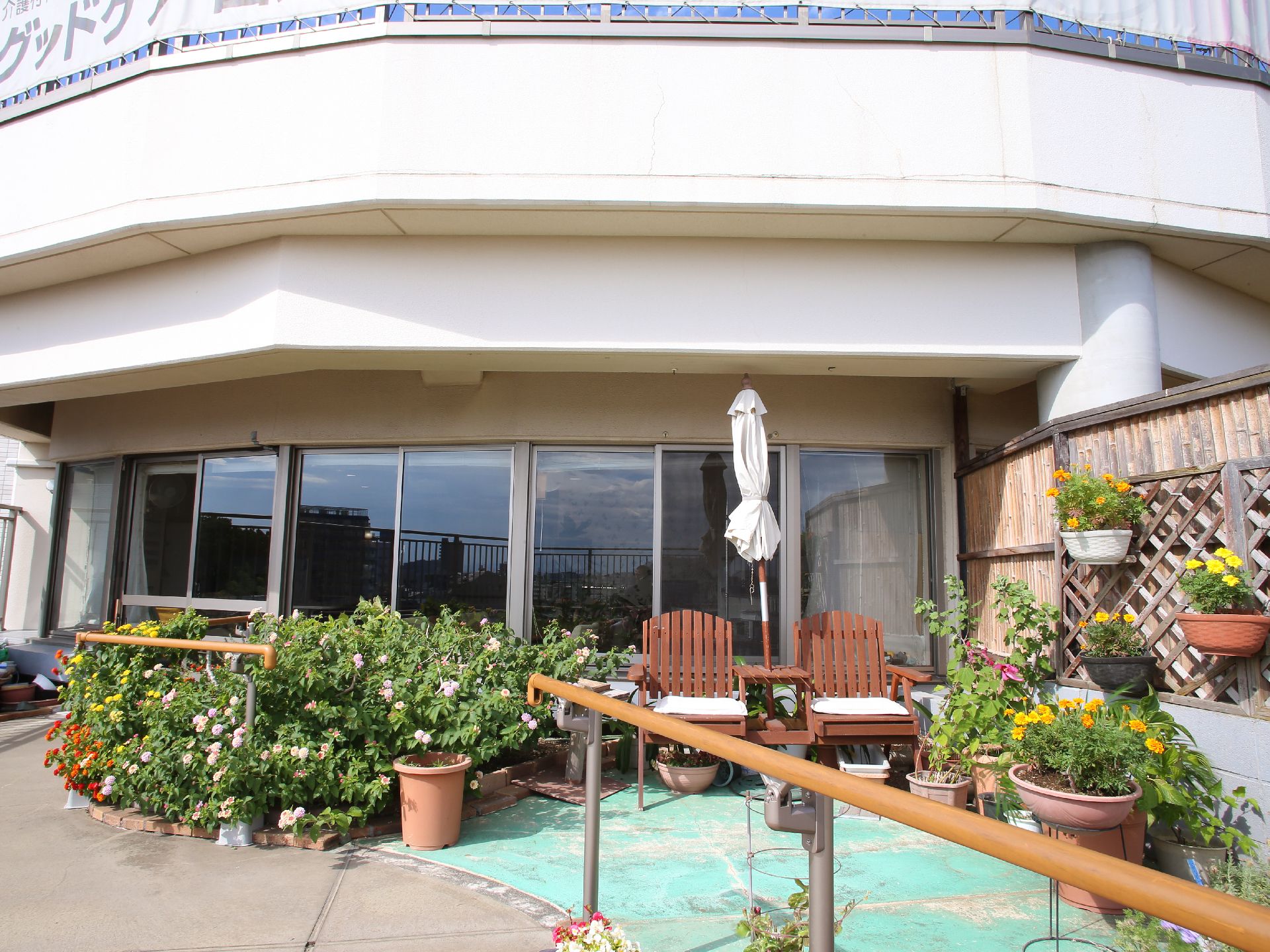 ウッドチェアー2脚とパラソルが置いてある。建物側は一面がガラス窓で少し円形になっている。ラティスにプランターが吊ってある。