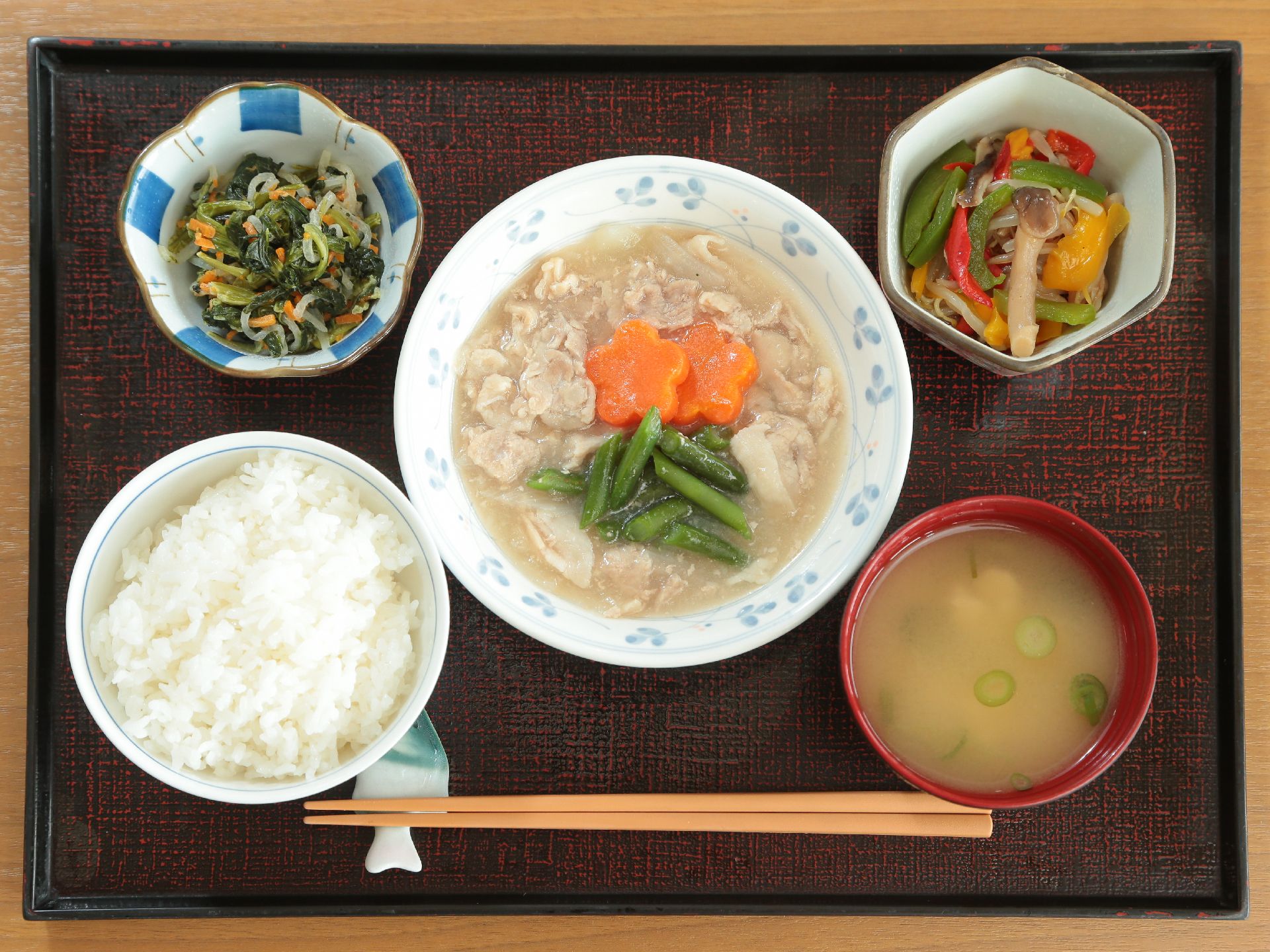 ご飯、汁物、メインおかず、小鉢2品、箸がトレーに載っている。ニンジンが飾り切りされている。彩り豊かである。
