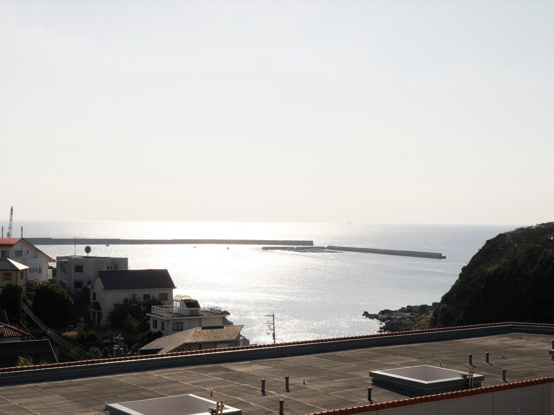 施設建物の前には広々とした海が広がっていて、日差しが反射している。海の手前には住宅と大きな岩場が見える。
