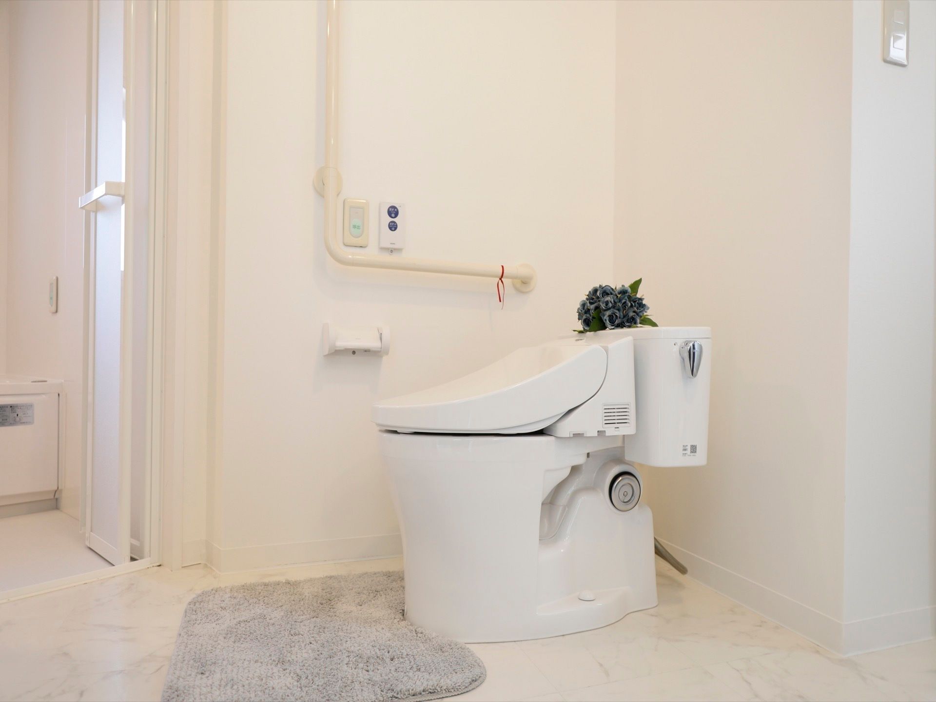 広々としたトイレ内は白で統一されており、明るく清潔感がある。便器の前にはグレーのマットが置かれている。