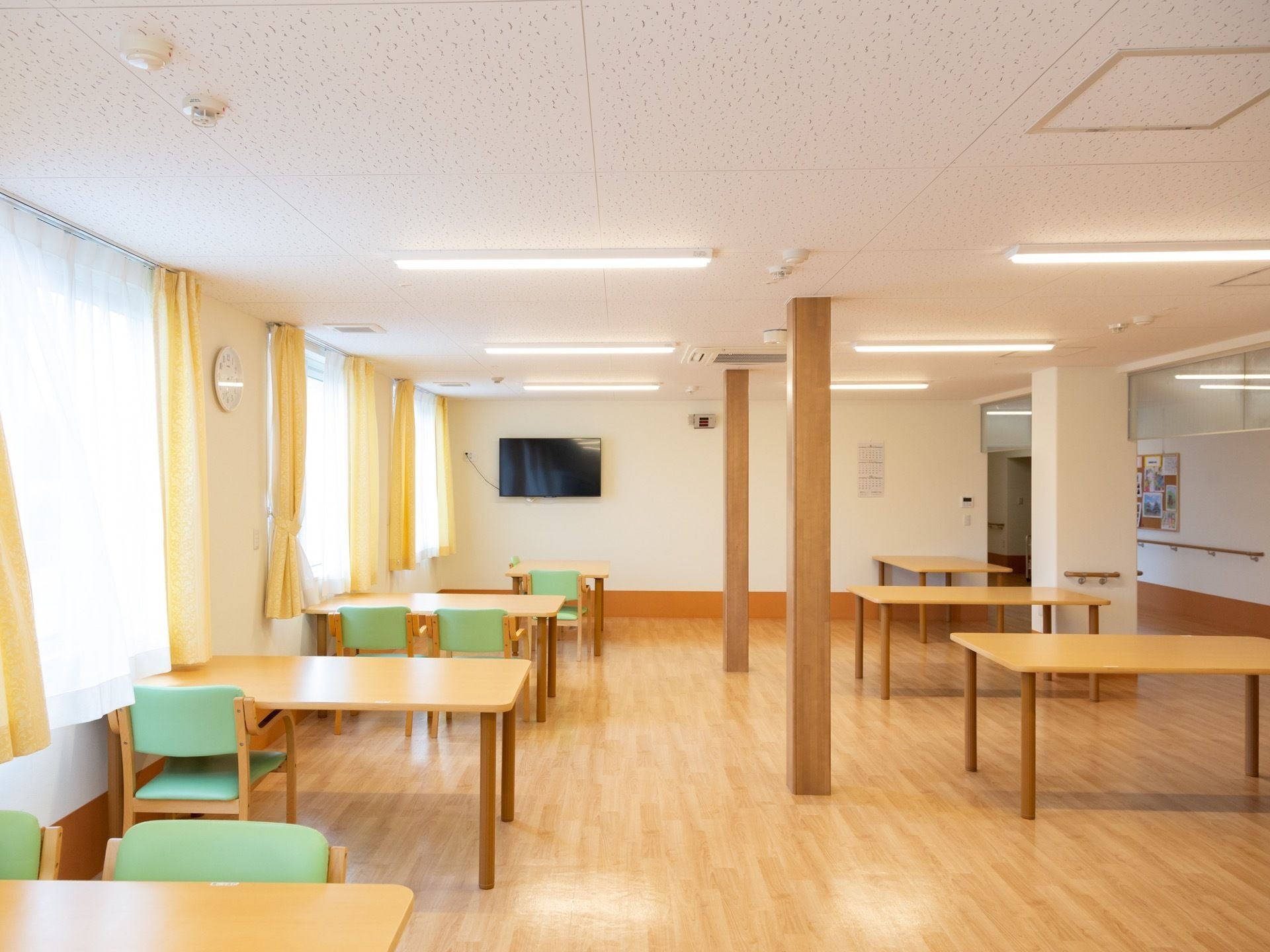 部屋の中にある柱を挟んで、ダイニングテーブルと緑色の椅子が置いてある。壁に黒い薄型テレビが掛けられている。