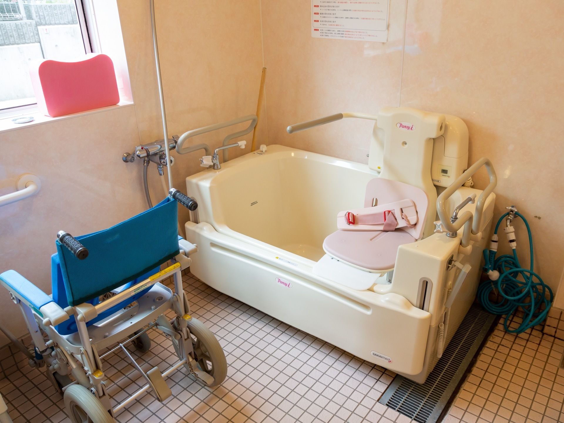 リフトの椅子がついた白い浴槽がある。洗い場に車輪がついた青いシャワー椅子が置いてあり、窓から外の景色が見える。