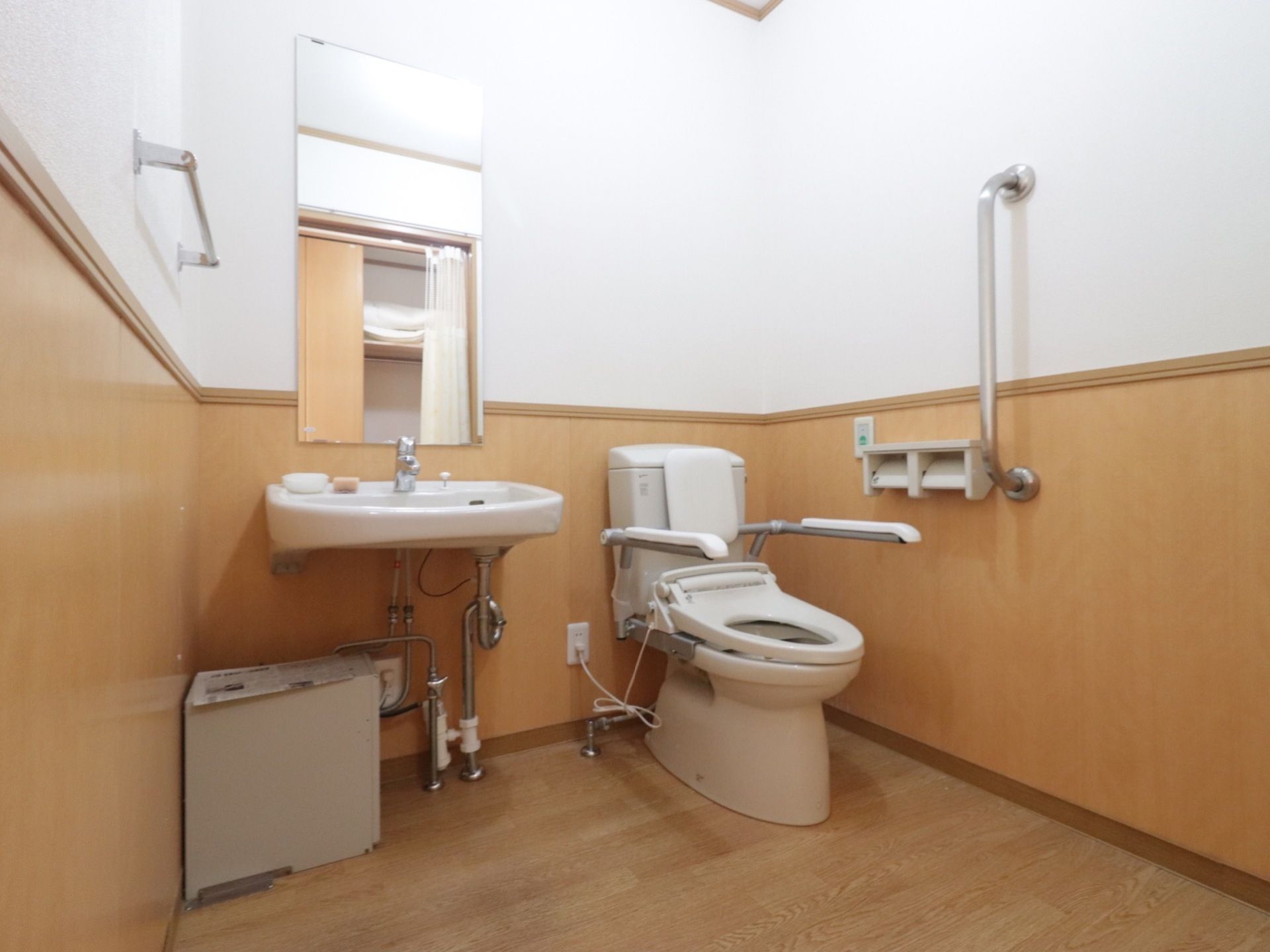 壁に手すりが設置されている。便器の近くには、緊急用のボタンがある。同じ空間に、洗面台、鏡、タオル掛けがある。