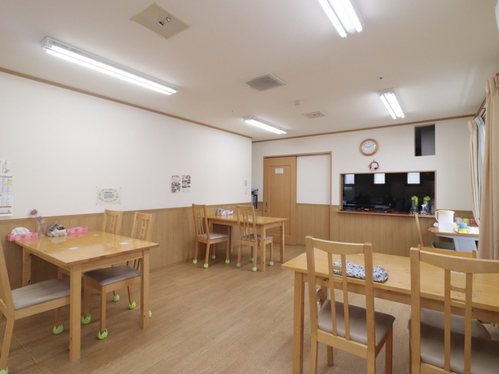 木製のテーブルと椅子が幾つも置かれている。椅子の脚には、布で作られたキャップがされている。ドアの近くにキッチンがある。
