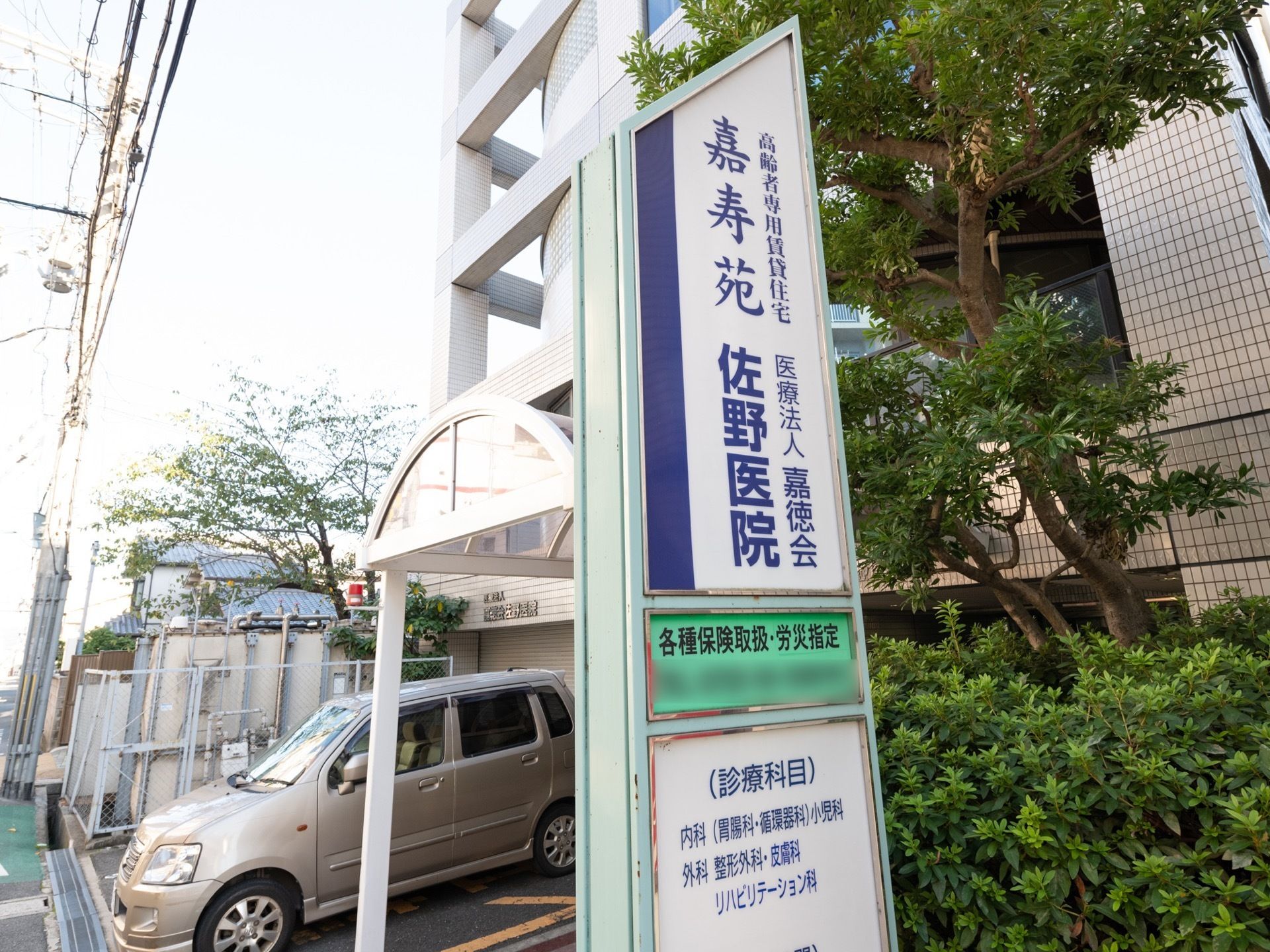 施設の入口前に施設名が書かれた看板が立てられている。併設の医院の診療科目が書かれ、その裏に入口がある。