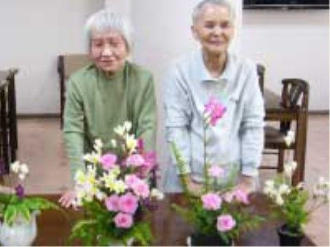 生花のレクリエーションの様子。テーブルに花を並べ、記念写真を撮る高齢女性２人。