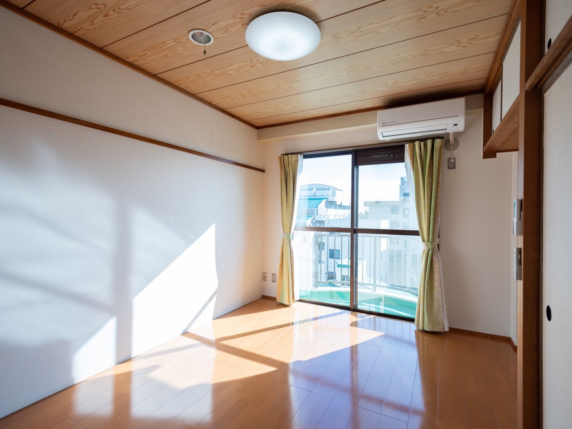 掃き出し窓から陽光が差し込む明るい居室である。床はフローリングとなっている。窓上にエアコンが設置されている。