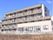 施設はベージュ色の外観で４階建てである。数多くの部屋の窓が並んでおり、それぞれベランダが付いている。施設の周りには植物が生えている。