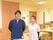 男性と女性の職員が各1名、横に並んでいる。男性のほうが若干、若く見える。両名とも笑顔で、背景はリビングルーム。