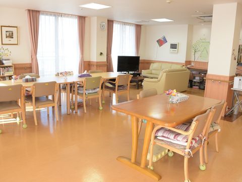 窓が大きく、明るいリビング。テーブルが3脚、4人掛けの椅子配列により並んでいる。左奥にテレビがあり、その手前にソファーが置かれている。