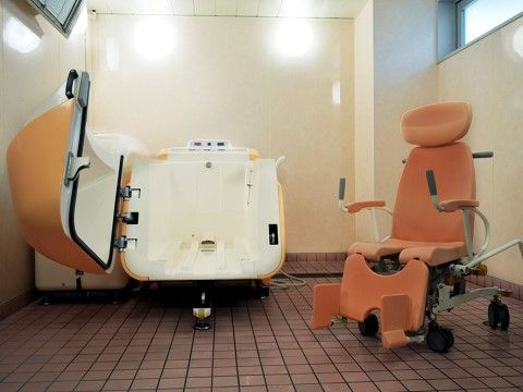 タイル張りの床の浴室に、オレンジ色の機械浴槽と椅子が置かれている。操作のしやすい機械浴用椅子で、安心して入浴できる。