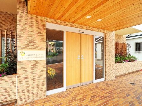 木目調の自動ドアと天井が温かみを感じる玄関。レンガの壁の両脇にはおしゃれなグリーンも植えられている。