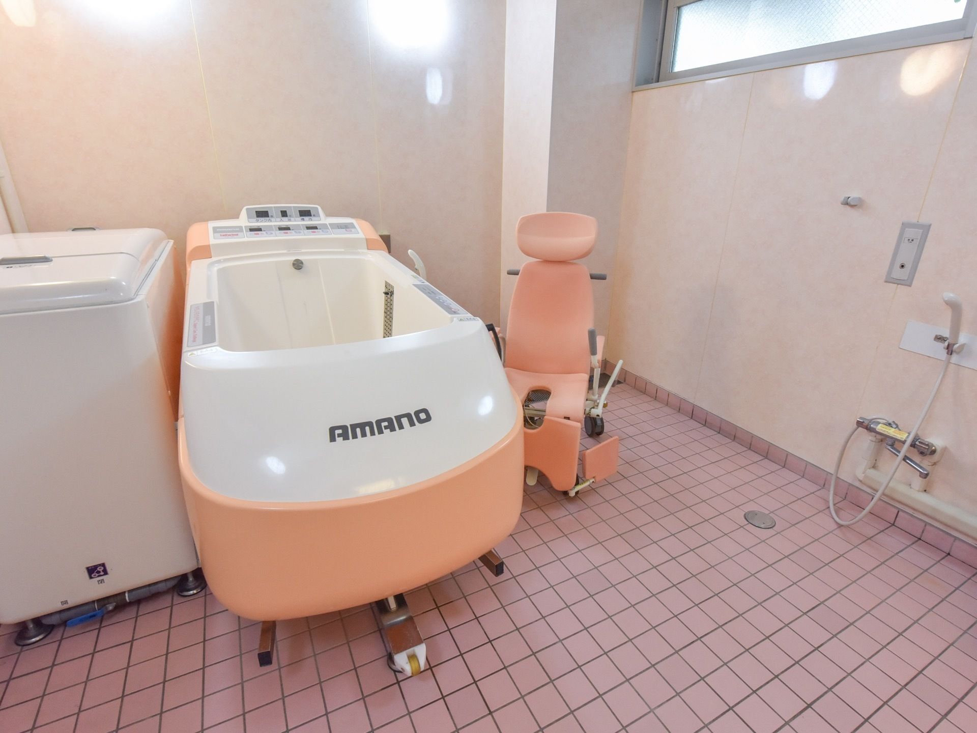 小窓が付いた個室の浴室に、腰を掛けたまま入浴することができる専用のシャワーチェアが付いた入浴器具が置かれている。