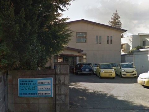 コンクリート壁に、青をベースとした施設名が書かれた四角い看板が設置されている。建物の前には複数台の車が停められている。
