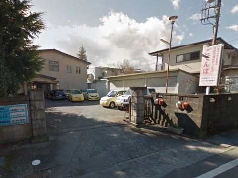 建物前にアスファルトで舗装された広い駐車スペースが設けられている。横にはシャッターの付いた大きな物置がある。