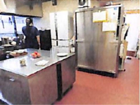 大きな調理台や冷蔵庫が置かれた広々とした厨房で、壁には換気扇が設置されている。女性スタッフが窓際に立ち、作業をしている。