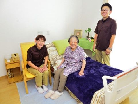 居室のベッドに腰かけている女性の入居者様と、その周りにいる男女１名ずつのスタッフ