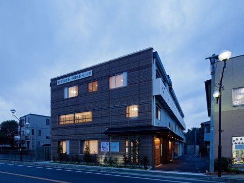 道路に面した部分の外壁に、施設名が記してある。１階には、壁際に樹木を並べて植えている。横にライン状に凹凸がある外装である。