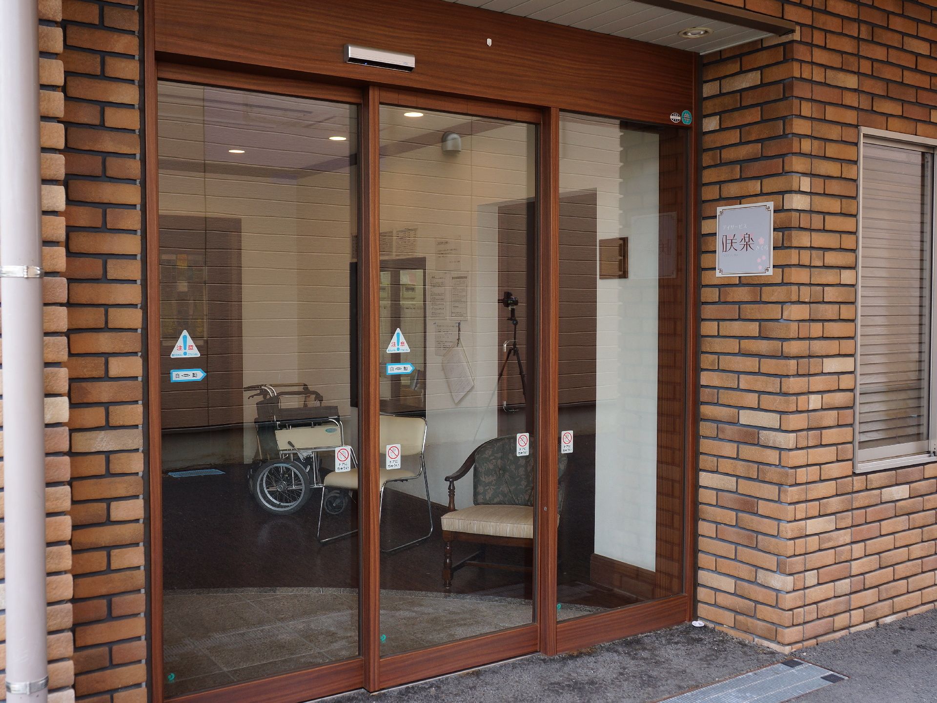 デイサービスの表札もついている玄関は、透明窓が3枚ついた自動ドアである。内部には、車椅子と椅子も用意されている。