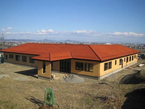 まだ造成したばかりの土地に、新しい建物が建っている。黄色い壁にオレンジの切妻屋根が設けられており、Ｌ字型の建物にひさし部分が飛び出ている。