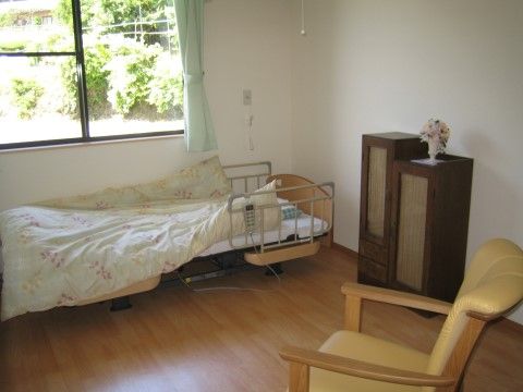 正面にある腰高窓の外には、敷地内の畑が見える。窓下には介護ベッドがあり、窓の横には非常用ブザーがある。
