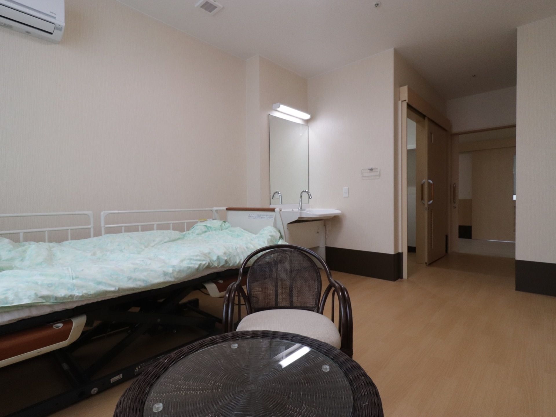白い壁紙の居室にはライムグリーンの布団が敷かれた介助用ベッドがある。洗面台と椅子、机、エアコンがある。