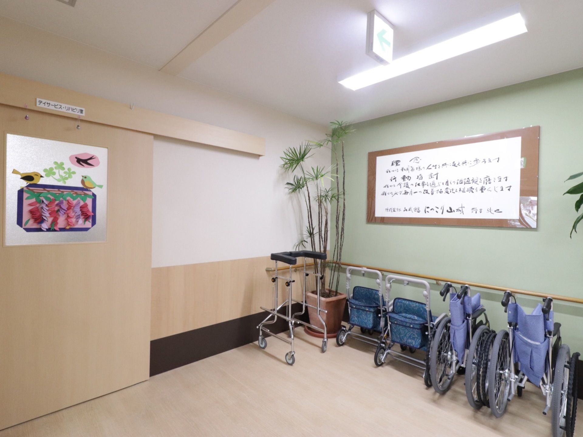 デイサービス、リハビリ室の入口である。壁に沿って車椅子や歩行補助器が並んでいる。壁には手すりがついている。